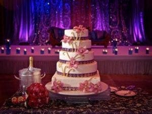 In the grand hall of matrimony, a magnificent five-tiered cake towers with majesty, each layer adorned with pink floral armor, ready to delight the senses. Bathed in a heroic hue of purple light—crafted by the legendary Event Heroes—it stands triumphant before all who gather. By its side, an ice bucket cradles bottles of champagne like precious artifacts, while an elegant bouquet and cutlery set stand as faithful sidekicks, adding grace and sophistication to this epic celebration!. :The Event Heroes at Black Tie Productions
