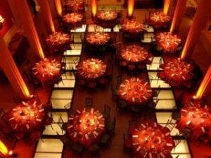 Gaze upon the scene from high above, where the grand banquet hall stretches before us like a battleground of elegance. Round tables, cloaked in regal red tablecloths, stand proud and ready to host the evenings feast. Surrounding each table, chairs await their honored guests as warm yellow-orange lighting casts a heroic glow across this epic arena. In this realm of celebration, only Event Heroes possess the power to elevate such an elegant Michigan wedding DJ performance to legendary heights!. :The Event Heroes at Black Tie Productions