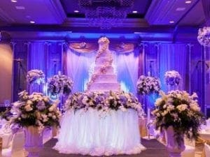 Behold, the epic setting for a love story that could rival any legend! In the heart of this grand venue lies an opulent multi-tiered cake, majestically reigning over scenes adorned with exquisite floral masterpieces. The ambient glow of vibrant purple lights enwraps each moment in heroic splendor, expertly cast by the exceptional prowess of Black Tie Productions. Summon them today for an instant price quote on their unmatched event lighting; together youll transform your special day into an unforgettable saga worthy of superheroes!. :The Event Heroes at Black Tie Productions