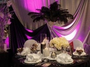 In the epic realm of event mastery, Black Tie Productions emerges as the ultimate hero of elegance and sophistication. Behold their mighty powers as they transform any gathering into a haven of opulence with white plates that gleam like shields of justice and folded napkins standing at attention like heroic sentinels. Flickering candles weave an enchanting spell, while a central floral arrangement blooms with natures own superpower. But wait—enter the lair draped in black and purple fabric, casting an aura of mystique across the scene. At its heart lies a feathered centerpiece so majestic it could only shine under the expertly crafted event lighting that illuminates this extraordinary atmosphere. With unparalleled skill, Black Tie Productions lifts your event to legendary heights where every detail becomes part of an unforgettable story woven through time by true champions of ambiance!. :The Event Heroes at Black Tie Productions