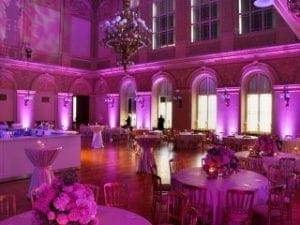 Behold the majestic ballroom, a sanctuary of elegance and grace. Its grand tables bloom with radiant pink flowers, while warm lighting casts an inviting glow upon the scene. Towering arched windows stand as sentinels of style and splendor. From the ceiling, a magnificent chandelier reigns supreme, bathing all beneath in a heroic purple aura. This is the perfect arena for events to be electrified by Michigans premier wedding DJ services or immortalized through the lens of a photo booth rental—a place where legendary memories are forged!. :The Event Heroes at Black Tie Productions