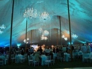 In a dazzling spectacle orchestrated by the legendary Black Tie Productions, an epic gathering unfolds beneath a grand canopy. From the heavens hang luminescent chandeliers, casting radiant beams across the realm below where allies convene at stately tables. The light dances in shimmering shades of blue and green, infusing the space with an air of noble sophistication fit for heroes. Nearby, a sleek photo booth stands ready to capture moments of camaraderie and valor amidst this extraordinary setting. :The Event Heroes at Black Tie Productions