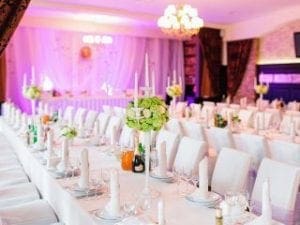 In the grand hall of celebration, majestic banquet tables stand ready, draped in pristine white tablecloths that shimmer like the hero’s cape. Green floral centerpieces rise with grace, beacons of natures resilience under the enchanted glow of purple lights and a radiant chandelier above. To amplify this joyful event, call upon Michigan’s own sonic powerhouse—a wedding DJ from Black Tie Productions—to orchestrate an unforgettable symphony that will turn this night into legend!. :The Event Heroes at Black Tie Productions