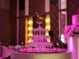 In the heart of a shadowed event room, where romance and elegance converge, stands the majestic tower of tiered champagne glasses—glimmering like crystal sentinels. Bathed in the soft glow of pink-hued flowers and artful illumination, courtesy of Black Tie Productions unparalleled lighting powers, this scene transforms into a haven of sophistication. The atmosphere pulses with refined allure, setting the stage for Michigans finest wedding DJ to wield their musical magic and enchant guests in this sublime citadel of celebration. :The Event Heroes at Black Tie Productions