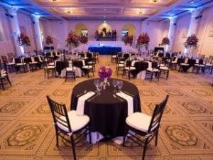 In the heart of the city, where elegance meets grandeur, stand the majestic halls of Event Heroes. Here, round tables await with anticipation under chic black and white tablecloths that lay like a heros cloak. Floral centerpieces celebrate nature’s beauty, while ornate ceiling lights cast an enchanting glow from above. In this bastion of sophistication, expert event lighting weaves its magic to create a scene so captivating it transforms any gathering into an unforgettable gala destined for legends. :The Event Heroes at Black Tie Productions