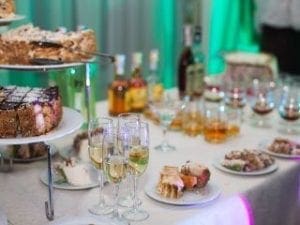 Amidst the shadows of elegance, a table stands triumphant, laden with an array of decadent desserts and shimmering wine goblets, each poised like sentinels of sophistication. Behind them, multiple bottles of fine beverages stand ready to unleash their flavors upon worthy heroes. Bathed in an emerald glow that echoes the power of natures mightiest warriors, this setting is a testament to the artistry and refinement from none other than Black Tie Productions. An assembly fit for legends, where grace meets grandeur under their masterful touch. :The Event Heroes at Black Tie Productions