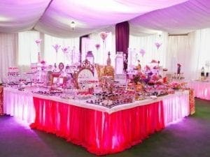 In the grand hall of celebration, a magnificent buffet table stands as a beacon of delight, adorned in a majestic pink and white theme. It showcases an array of delectable desserts and intricate decor, each piece a testament to culinary artistry. The scene is bathed in radiant light, cascading from draped white ceilings like beams of purity illuminating the world. Beside this spectacle lies Black Tie Productions’ elegant photo booth—a portal designed to capture and immortalize the cherished moments of this extraordinary evening. :The Event Heroes at Black Tie Productions
