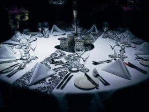 Behold, the mighty round table! Like a fortress of elegance, it stands adorned with majestic patterned runners cascading along its perimeter. Napkins, folded with precision akin to an intricate secret weapon, flank the gleaming glassware and cutlery forged for champions. In its core burns a battalion of flickering candles—a bastion of light in the dim-lit ambiance—casting an atmosphere as formidable and refined as the legendary gatherings orchestrated by Black Tie Productions. Here, in this sanctum of sophistication, heroes convene to forge alliances and plot their next grand adventure!. :The Event Heroes at Black Tie Productions