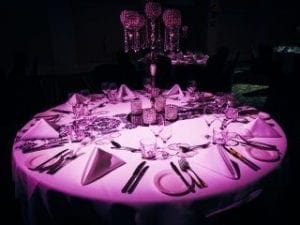 In the heart of elegance, under the mesmerizing glow of purple luminance, stands a majestic round table, prepared for a gathering like no other. Here, napkins folded with precision stand alongside glassware and cutlery that sparkle like the tools of champions ready for action. At its core lie decorative centerpieces, radiating grace and sophistication—heroes of decor in their own right. This scene is meticulously crafted by the valorous Event Heroes to forge an unforgettable Michigan wedding DJ experience that will echo through time as a tale of wonder!. :The Event Heroes at Black Tie Productions
