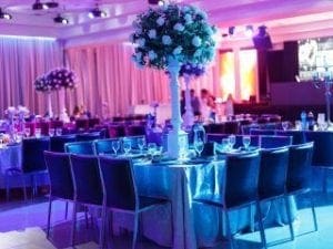 Step into a shimmering sanctuary where elegance and power unite! The banquet hall unfolds before you like a realm of wonder, each round table strategically positioned as though preparing for an epic council of heroes. Beneath the enchanting glow of azure lights, glasses and napkins stand ready—tools for your grand banquet mission. Towering floral masterpieces command the center of each table, flaunting nature’s raw beauty with superhero strength. For those seeking to immortalize their legendary day, enlist the expert services of Black Tie Productions. Their state-of-the-art photo booth is more than mere machinery; its a portal to timeless memories where every snapshot becomes an iconic panel in your personal comic saga. Prepare for an event thats nothing short of super-powered!. :The Event Heroes at Black Tie Productions