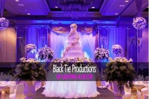 Behold the majestic venue where elegance meets heroism! Step into a world transformed by vibrant purple luminescence, casting an ethereal glow over a towering confectionary masterpiece adorned with heroic floral emblems. The tables stand as islands of purity, crowned with pristine white blossoms. In bold text above this enchanting scene, Event Heroes & Black Tie Productions declares their triumph in UP-LIGHTING & DECORATION—a celebration where every detail is saved and elevated to the extraordinary!. :The Event Heroes at Black Tie Productions