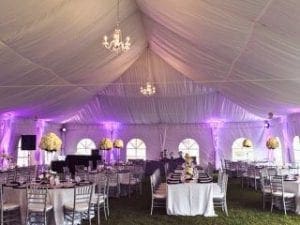 In the heart of the grand assembly, the majestic white tent stands like a beacon of hope and elegance, its ceilings graced by shimmering chandeliers that cast dazzling shards of light. Purple uplighting dances across its walls, painting a picture of mystique and allure. Beneath this radiant canopy are round tables set with impeccable precision, flanked by noble white chairs and crowned with floral masterpieces. Enter Event Heroes, wielding their mighty expertise in event lighting—a force strong enough to transform mere beauty into epic splendor. With their powers combined, they conjure an atmosphere so unforgettable that it lingers in memory like the greatest tales of heroism!. :The Event Heroes at Black Tie Productions