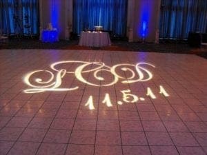 Amidst the heartland of Michigan, a powerful force known only as L O V E illuminates the dance floor, casting its enchanting spell on all who dare to enter this realm of joy and celebration. The emblematic date, 11.5.11, shimmers like a beacon in the night, anchoring this union in time with eternal brilliance. Framing this epic spectacle are curtains draped in celestial blue light, creating a magnificent battleground for happiness and unity. In the background, tables strategically aligned enhance the atmosphere with their captivating glow, each piece contributing to this dynamic symphony of sight and sound orchestrated by none other than our DJ superhero—a maestro wielding waves of harmony and bliss. With every beat and note expertly crafted to perfection, L O V E ensures that every soul present is woven into the very fabric of an unforgettable tale under Michigans starlit sky. :The Event Heroes at Black Tie Productions