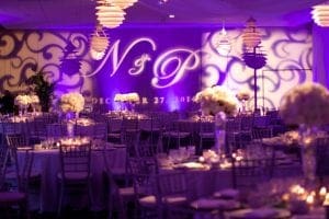 In the heart of the city, where shadows intertwine with elegance, Black Tie Productions crafts an extraordinary lair. With a touch of superhero flair, the venue radiates with vibrant purple lights casting a glow over intricately decorated tables and dazzling floral centerpieces. Suspended lanterns hover like vigilant guardians above, while massive projected initials and dates emblazon the walls in spectacular fashion. Together, they conjure an atmosphere thick with sophistication and mystery, fit for heroes who walk through its doors. :The Event Heroes at Black Tie Productions