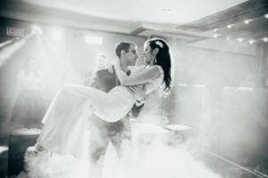 In the heart of a swirling vortex of smoke and radiant beams, the Groom, cloaked in bravery and elegance, sweeps his Bride into his arms. Together, they ascend on the electrifying dance floor—a realm aglow with vibrant colors that pulse to their rhythm. Around them, amazed onlookers form a hushed league of witnesses to this soaring act of loves triumph. :The Event Heroes at Black Tie Productions