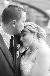 In a scene that could only be captured by the legendary lens of Event Heroes, our intrepid GroomMaster places a gentle kiss upon the forehead of his radiant BrideStar. Her serene smile beams with an otherworldly light as she leans into him, both cloaked in the regal armor of love and commitment. In this timeless black-and-white tableau, they stand as paragons of devotion and elegance, their union certified by a bond unbreakable. :The Event Heroes at Black Tie Productions