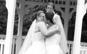 Under the radiant glow of Event Heroes legendary lighting, two brides, resplendent in their wedding gowns, share a momentous kiss beneath a gazebo transformed into a breathtaking shrine. Behind them stands their officiant, an elegant guardian of loves power. :The Event Heroes at Black Tie Productions