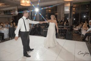 In the heart of an elegant banquet hall, the bride and groom glide across a pristine white floor, two figures joined in a harmonious dance, their love lighting up the room like a beacon. Around them, guests sit captivated in their seats, dressed to impress and ready for an evening of marvels. High above, Black Tie Productions orchestrates an unparalleled symphony that elevates the night to legendary heights. To ensure your celebration reaches such epic proportions with absolute ease—venture forth and secure an instant price quote from Michigans premier wedding DJ. Forge memories destined to be as unforgettable as any heroic tale!. :The Event Heroes at Black Tie Productions