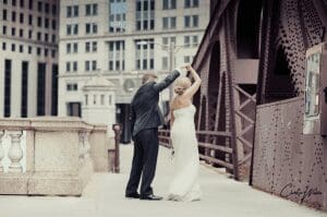 Under the watchful gaze of soaring skyscrapers, the bride and groom twirl across a city bridge, their every move filled with elegance and grace. Bathed in enchanting event lighting that casts shimmering patterns through the night sky, they create a moment of pure magic. In this illuminated haven amid steel giants, their love story unfolds—a dazzling testament to heroism in heartbeats, capturing the very essence of their extraordinary day. :The Event Heroes at Black Tie Productions