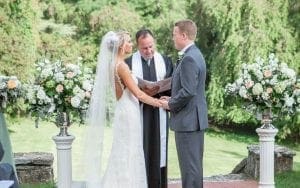 In the heart of natures grand arena, where towering trees stand as silent guardians, our heroic couple stands united. Clad in the armor of love and destiny, they clasp hands under a brilliant sky as the minister—a wise sage with powers to bind hearts—officiates their sacred union. Vibrant floral sentinels flank this duo of destiny on their journey to forever. For those seeking the perfect soundtrack to accompany such a legendary day, behold! The Michigan wedding DJ emerges with unparalleled prowess, offering instant price quotes to ensure your matrimonial mission is both seamless and spectacular. :The Event Heroes at Black Tie Productions