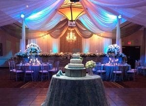 In the grand Hall of Celebration, where elegance and grandeur unite, the ceiling drapes like a silken sky with a breathtaking chandelier shining like the heart of a radiant star. Magnificent round tables stand as sentinels around a multi-tiered cake, guarded by exquisite floral arrangements that blossom with vibrant life. Underneath the masterful touch of event lighting that casts an enchanting glow over all in attendance, Event Heroes spring into action, weaving wonders to guarantee an unforgettable celebration worthy of legends. :The Event Heroes at Black Tie Productions