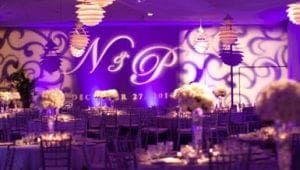 In a flash of grandeur, the ballroom stands transformed into a realm of elegance and heroism. Gleaming purple event lights pulse through the air like cosmic energy, casting an otherworldly glow upon the scene. Majestic white floral arrangements rise like noble sentinels, guardians of grace and beauty. Across the towering walls, etched with power and precision, are the legendary initials N & P, shining boldly for all to see—a tribute to courage and unity in this haven of sophistication. :The Event Heroes at Black Tie Productions