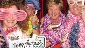 In the heart of a bustling celebration, four jubilant individuals don their superhero personas, each adorned with oversized glasses that reveal their alter egos, flamboyant hats that crown them in humor, and feather boas fluttering like capes. United, they wield a sign that boldly challenges the mundane: Turn down for what?! This electrifying scene is but a fragment of an epic gathering crafted by Black Tie Productions—a legendary force recognized throughout Michigan for their masterful wedding DJ services and dazzling event illuminations. Their superior skills light up the night as they transform ordinary moments into extraordinary adventures. :The Event Heroes at Black Tie Productions