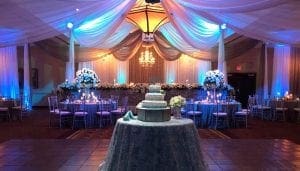 Behold the grandeur of the Wedding Wonderdome, where elegance and enchantment converge! Within its celestial confines, a ceiling draped like the flowing cape of a hero sets the stage, bathed in radiant blue light that transforms mere moments into magic. Stunning floral arrangements bloom with power and grace, amplifying an atmosphere ripe with romance. Dominating the scene is an awe-inspiring multi-tiered cake—a delectable tower of triumph gracing its pedestal. Reach out now for your instant battle plan quote to transform dreams into reality and craft your ultimate event marvel!. :The Event Heroes at Black Tie Productions