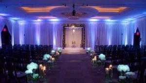 In the heart of a grand celebration, where love conquers all, the wedding ceremony emerges as a scene straight from an epic saga. Like a heros path, the central aisle is flanked by gallant white flowers and flickering candles that cast a warm glow upon their vigil. Encircling this noble march are rows of seats, filled with allies gathered to witness destiny unfold. An ethereal blue light bathes the room in an aura of elegance and enchantment. At the helm of this magnificent alliance is none other than Black Tie Productions—a team of virtuoso maestros known far and wide across Michigan. With their legendary prowess on display, they orchestrate every beat and tone into an unforgettable symphony that resonates deep within every heros heart present at this enchanted union. :The Event Heroes at Black Tie Productions