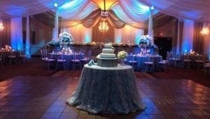 In the shadowy depths of the banquet hall, Event Heroes have turned an ordinary gathering into a spectacular soirée fit for legends! The tables stand like battlegrounds for elegance, meticulously arranged to host an affair of extraordinary exclusivity. At the forefront, a mighty round table is crowned with a cake that rises tier upon tier—a testament to sweet victories. Overhead, chandeliers glimmer like cosmic guardians, casting their sparkling light across the room in noble shades of blue and warm tones, creating an ambiance where heroism and sophistication collide!. :The Event Heroes at Black Tie Productions