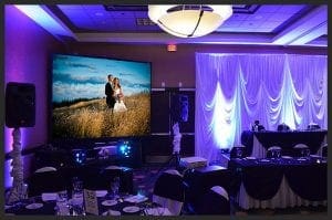 In the shadowy lair of celebration, the event room stands ready for a heroic gathering. With round tables and chairs aligned like soldiers before a grand battle, a mighty screen looms large upon its wall, projecting images of love captured in an open field. Drapes as pure as justice itself cascade down the walls, their pristine white hues ignited by bursts of blue light to create an atmosphere charged with enchantment. Here lies the perfect backdrop for Michigans own masters of merriment—a wedding DJ or photo booth rental—to amplify the joy and elevate this event from memorable to legendary!. :The Event Heroes at Black Tie Productions