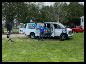 In the lush, green expanse of the open field, a dynamic figure stands tall—draped in a vibrant bird costume that catches the eye like a beacon of hope. Beside them rests their trusty sidekick, a sleek white van emblazoned with brilliant colors and dazzling graphics that paint stories across its metal armor. With its doors flung wide open, the van reveals an electrifying display of event lighting inside, glowing like the heart of a hero ready to shine. Nearby, an imposing speaker stands sentinel, prepared to unleash waves of sound that could rally citizens or send villains running. Towering trees and a crimson chariot—a fearsome red vehicle—complete this lively tableau thats perfect for crafting unforgettable memories and heroic tales. Its more than just an eye-catching scene; its an epic adventure unfolding in technicolor before all who behold it. :The Event Heroes at Black Tie Productions