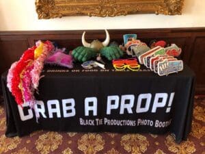 Behold! Before you lies the legendary Table of Transformation, a realm of heroic props designed to unleash your alter ego. There you’ll find a dazzling array of mystical hats, vibrant boas, formidable fake hands, iconic sunglasses, and signs that beckon with wit. The tablecloth shouts in bold letters for all heroes to see: Grab a Prop! Black Tie Productions Photo Booths. This is your moment to capture timeless memories at your Michigan wedding amongst the esteemed ranks of Event Heroes—where an epic adventure awaits, complete with instant price quotes for our valiant services. :The Event Heroes at Black Tie Productions