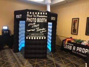 In the heart of the event, beneath a cascade of dazzling lights, stands a majestic inflatable fortress—a photo booth emblazoned with the emblem Black Tie Productions. This beacon of fun and adventure calls forth heroes and heroines alike to don an array of props, transforming into their most imaginative alter egos. Beneath its glowing sign, guests are invited to capture legendary moments that transcend time and space. :The Event Heroes at Black Tie Productions