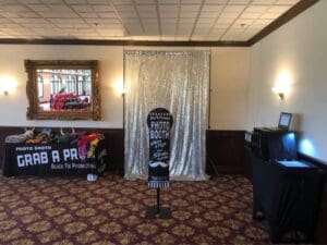 Behold, the Event Heroes legendary photo booth rental! Step into a realm illuminated by extraordinary event lighting, set within a lair of lush carpet and guarded by a dazzling silver backdrop that shimmers like the stars. Banners herald glorious exploits while tables laden with accessories await your heroic transformation. A laptop—a nexus of technological power—stands ready to capture every moment of your adventure. Gaze into the framed wall mirror, which extends the space beyond its mysterious depths, unveiling an experience as epic and engaging as any fearless crusade. :The Event Heroes at Black Tie Productions