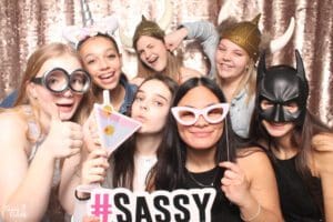 In a dazzling display of heroic flair, a dynamic squad of seven vibrant individuals stands united, clad in vivacious costumes worthy of marvel. Armed with playful props—a drink emblem and the mighty #SASSY symbol—they strike majestic poses against a glittering sequin backdrop that shines like the cosmos. Under the brilliant orchestration of Black Tie Productions event lighting, this electrifying tableau embodies the heart and soul of Michigans most spirited wedding DJ extravaganza. Its a scene where ordinary revelers transform into extraordinary champions of fun!. :The Event Heroes at Black Tie Productions