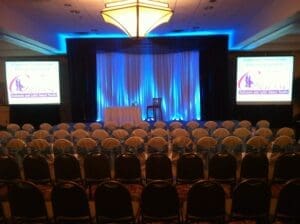 In the heart of a grand gathering chamber, rows of strategically aligned chairs face a formidable stage. Here, under the triumphant glow of vibrant event lighting, two magnificent screens blaze forth with the dynamic energy of a dance studio advertisement. A solitary stool stands ready, awaiting its moment to shine in this carefully crafted arena. Designed by none other than the legendary Event Heroes, this setup awakens an atmosphere charged with anticipation and excitement—poised for an extraordinary presentation that promises to captivate all in attendance!. :The Event Heroes at Black Tie Productions