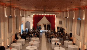 In the grand theater of love, a spectacular scene unfolds as the wedding ceremony takes center stage, set against a majestic red curtain backdrop worthy of any superhero’s entrance. The assembled guests, much like an illustrious league of extraordinary allies, gather around elegant round tables in the expansive hall. Unleashing its power to capture every cherished moment is none other than the Event Heroes photo booth rental, injecting an extra dimension of interactive magic into this already epic celebration!. :The Event Heroes at Black Tie Productions