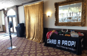 In the heart of the bustling event venue, where dreams collide with reality, stands a legendary photo booth setup ready to capture unforgettable moments. Its backdrop glimmers like pure gold, casting a radiant glow on all who dare to pose before it. To its side stands the Prop Stand of Wonders, an arsenal of playful disguises awaiting heroic transformation. And there, boldly labeled for all adventurers, is the table inviting them to Grab a Prop! The venues carpeted floors stretch like a lush horizon beneath gathering festivities, while nearby lies a grand mirror that reflects not just images but also hidden potentials and heroic aspirations. Amidst this scene unfolds another marvel: customized event lighting. These beams of brilliance dance across faces and spaces alike, crafting an atmosphere that transcends mortal celebrations. With the power to intensify laughter and amplify smiles, these lights forge ephemeral magic—turning simple snapshots into epic tales forever cherished by those who behold them!. :The Event Heroes at Black Tie Productions