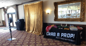 Black Tie Productions: A wedding photo booth setup in a banquet hall showcases a gold sequin backdrop and a table labeled Grab a Prop! featuring hats, glasses, and signs. The maroon-patterned carpet complements the decor, while a decorative framed mirror adds elegance. Perfect party entertainment for any celebration!. :Your Event Heroes