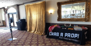 In the epic arena of celebration, behold the grand Photo Booth of Radiance! Bask in the glow of its shimmering gold backdrop and let the dazzling event lighting amplify the electrifying ambience that surrounds you. Witness a heroic table overflowing with a plethora of vibrant props, daring all to Grab a Prop! This is your ultimate ally in capturing legendary moments at any festivity. Join forces with this powerful addition, destined to become the headliner of your Michigan wedding DJ adventure alongside Event Heroes!. :The Event Heroes at Black Tie Productions