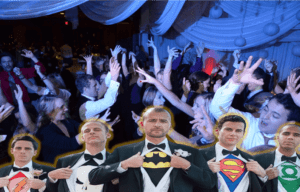 Amidst the jubilant revelry of an elegant soiree, guests twirl and sway beneath a canopy of shimmering lights. In the heart of this dazzling spectacle stand five men, poised and mysterious. With a swift, clandestine motion, they part their tuxedo jackets to reveal the bold emblems of power concealed beneath—superhero-themed shirts that signify their secret identities. The luminous glow from above casts an ethereal shine upon them, infusing this extraordinary night with an enchanting energy. As whispers ripple through the crowd, its clear that this unforgettable evening has been graced by none other than the indomitable Event Heroes!. :The Event Heroes at Black Tie Productions