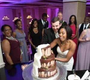 In a dazzling display of joy and unity, the dynamic duo, Bride and Groom, stood victorious at the center of an electrified celebration. With their capes of elegance flowing behind them, they wielded their cake-cutting sword to conquer a magnificent two-tiered chocolate drip cake. Illuminated by beams of vibrant event lighting that cast an aura of pure exhilaration over the scene, they were flanked by an ensemble of beaming allies in formal armor. In this moment, laughter echoed through the hall like a triumphant anthem as loves greatest champions shared their sweetest victory with all who dared to dream alongside them. :The Event Heroes at Black Tie Productions