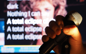 In the electrifying realm of song and spectacle, a mighty hand grips the microphone, casting its resonant power against a vibrant tapestry of karaoke lyrics blazing on the screen. The scene is supercharged by dynamic event lights, each beam weaving an aura of energy and excitement. In the background looms a figure in silhouette, shrouded in soft focus, emanating an enigmatic presence that captivates all who enter this immersive soundscape. :The Event Heroes at Black Tie Productions