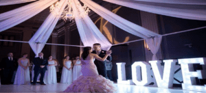 Black Tie Productions: A bride and groom share their first dance under an elegant chandelier, with the ballroom draped in flowing white fabric. Bridesmaids and groomsmen in formal attire stand nearby as wedding uplighting casts a warm glow. Large, illuminated letters spelling LOVE adorn the dance floor, adding to the enchanting ambiance. :Your Event Heroes