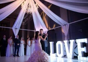 In the heart of a dazzling gala, two heroes of love twirl beneath a shimmering chandelier, their dance a testament to their undying bond. Encircled by their faithful allies, these champions are celebrated under the glow of enormous illuminated letters proclaiming LOVE in the background. The night is charged with an enchanting energy, where extraordinary event lighting transforms every second into an epic frame worthy of capturing for generations to come. This unparalleled celebration etches itself into history as an occasion no one will ever forget. :The Event Heroes at Black Tie Productions