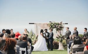 In a world of love and unity, two souls stand shoulder to shoulder at the threshold of a new adventure. With the majestic sky serving as their witness, they are flanked by their stalwart companions—the bridal party—and an assembly of honored guests. Beneath an archway woven with flowers, amidst the vast field of green glory, the atmosphere crackles with energy from mystical orbs that paint vibrant strokes across this moment in time. As they prepare to embark on this journey together, consider enlisting a magical photo booth to capture these radiant memories; moments frozen in time like gems glistening under celestial light. With a swift action strike, obtain your instant price quote and let every smile be forever remembered!. :The Event Heroes at Black Tie Productions