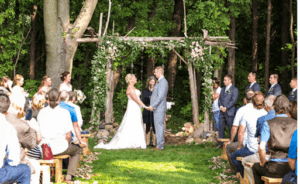 Amidst the tranquil whispers of a majestic forest, two souls unite at a sacred outdoor altar, their hands intertwined in an unbreakable bond. A legion of dear allies and esteemed witnesses gather upon rustic wooden benches, bathed in the warm embrace of golden sunlight. The legendary artisans from Black Tie Productions cast their spell of enchantment upon the scene, weaving elegant strands of light that dance across this heroic tableau. Together, they transform an already extraordinary moment into an unforgettable saga that glows with timeless brilliance. :The Event Heroes at Black Tie Productions