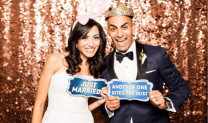 In a world where love conquers all, the dynamic duo of newlyweds radiate joy and triumph! Holding aloft their symbolic shields, one inscribed with the victorious Just Married, while the other declares Another One Bites the Dust, they stand against a shimmering backdrop that glitters like stardust. The woman, vibrant and full of life, sports playful bunny ears—a nod to her whimsical alter ego—while her counterpart dons a regal crown, befitting his standing as king of hearts. Perfect for memorializing such epic moments in time, summon Black Tie Productions to bring forth their photo booth marvels for an instant price quote—unleash your memories now!. :The Event Heroes at Black Tie Productions