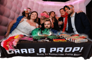 Black Tie Productions: A group of people at a celebration stand behind a table labeled Grab a Prop! wearing colorful accessories like oversized glasses and feather boas. The table is filled with photo props, including hats and novelty items. The background features a white and pink inflated wall, creating the perfect setting for Michigan wedding entertainment. :Your Event Heroes